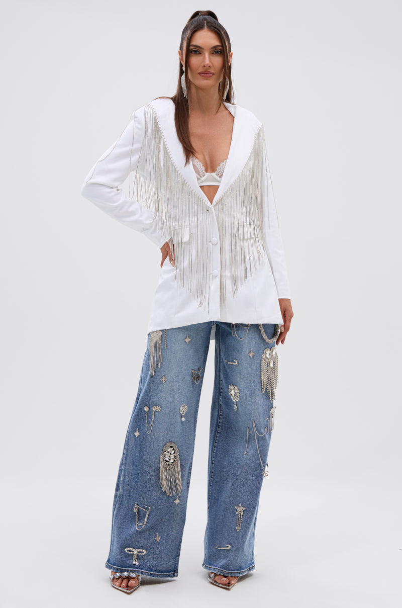 A woman faces forward against a white background, wearing the DRIPPY RHINESTONE FRINGE BLAZER IN WHITE over a visible bralette, wide-legged blue jeans with metallic embellishments, and open-toe heels. 7