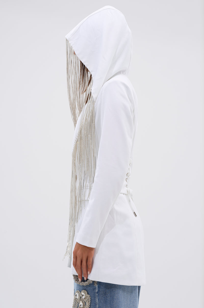 A person wears the DRIPPY RHINESTONE FRINGE BLAZER IN WHITE, featuring long rhinestone fringe covering their face, paired with light blue embroidered jeans. They're seen from the side against a plain white background. 8