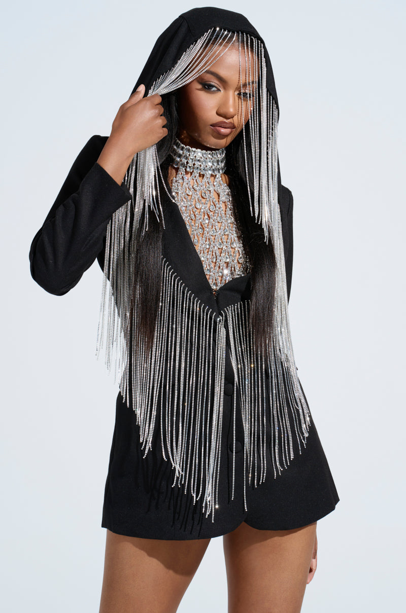A woman poses confidently in the DRIPPY RHINESTONE FRINGE BLAZER over a black hooded jacket with sparkling silver accents. Her polished, long hair flows through the fringe, while a plunging neckline adds allure against the plain white backdrop.