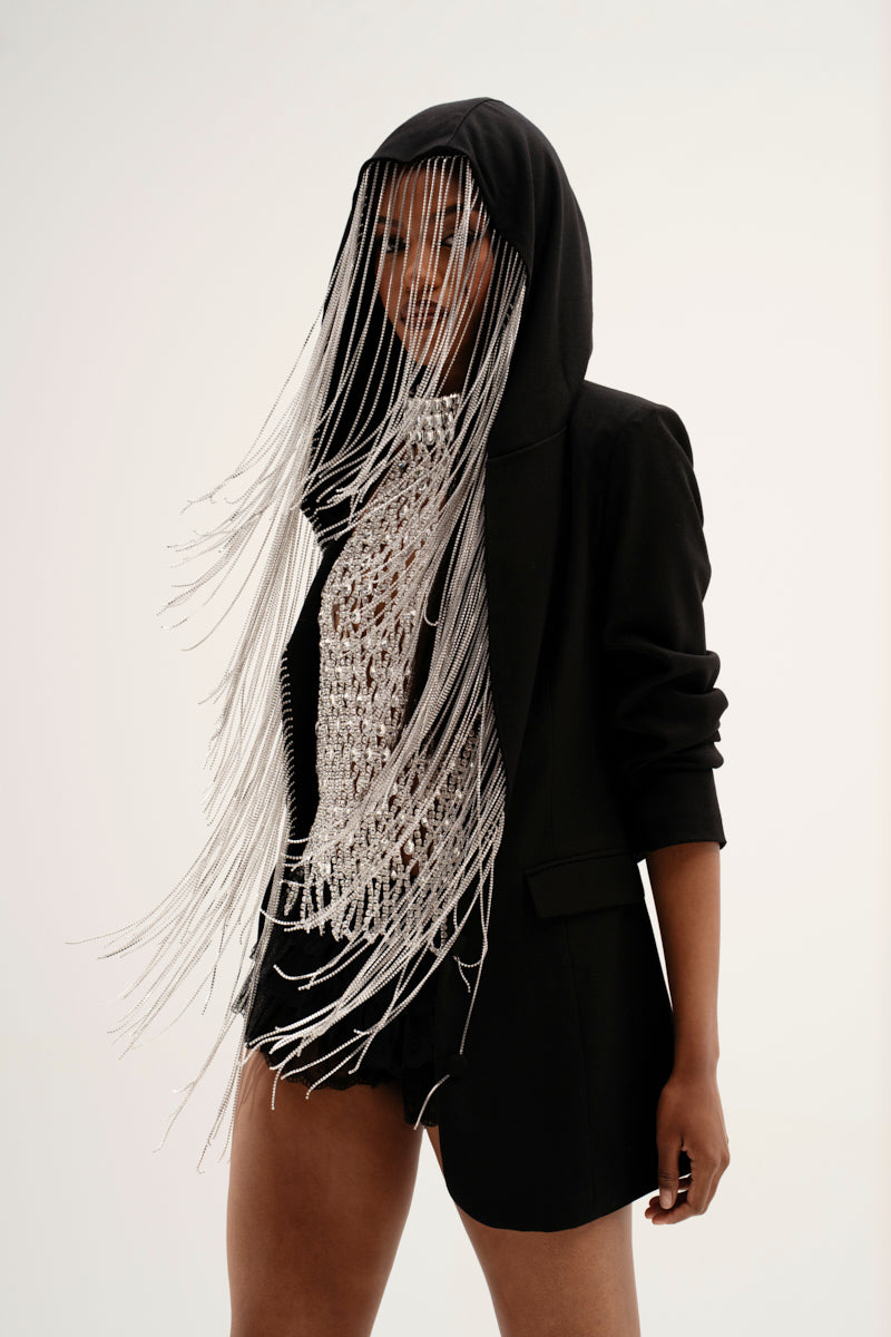 Clad in a stylish black hoodie and the DRIPPY RHINESTONE FRINGE BLAZER, complete with long silver strands concealing their face, they showcase a sparkling textured top with a plunging neckline and black shorts against a plain white backdrop that highlights the ensemble.