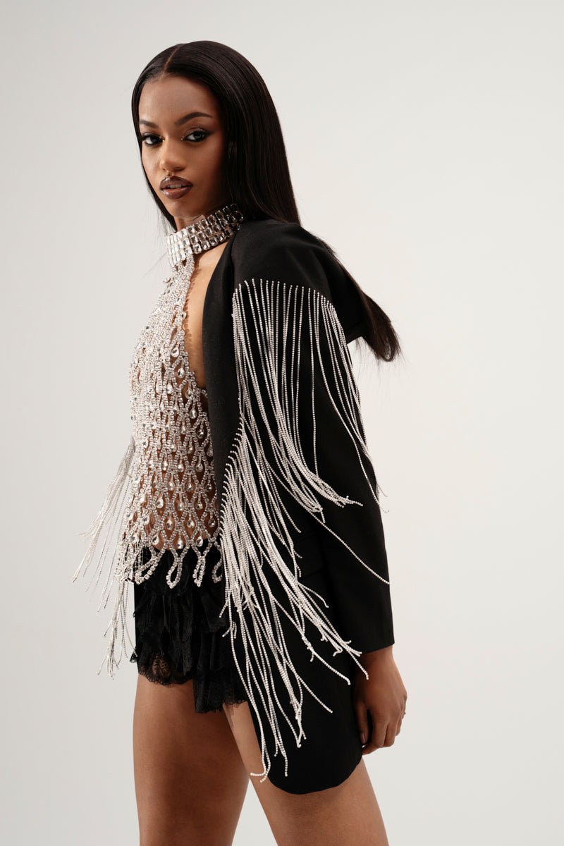 A person models the DRIPPY RHINESTONE FRINGE BLAZER, a stylish black piece featuring a lace-up corset detail with metallic fringe and a mesh, beaded top. The look is complemented by a matching choker, set against a plain background that highlights this fashionable ensemble.