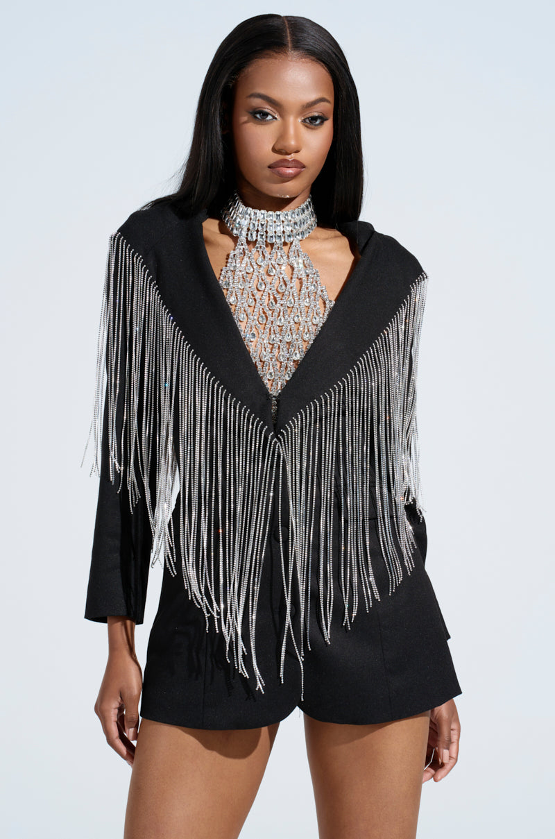 A woman confidently poses against a light background, showcasing her fashion-forward style in the DRIPPY RHINESTONE FRINGE BLAZER and a shimmering beaded halter top. With long, straight hair and an assertive expression, she embodies chic elegance.