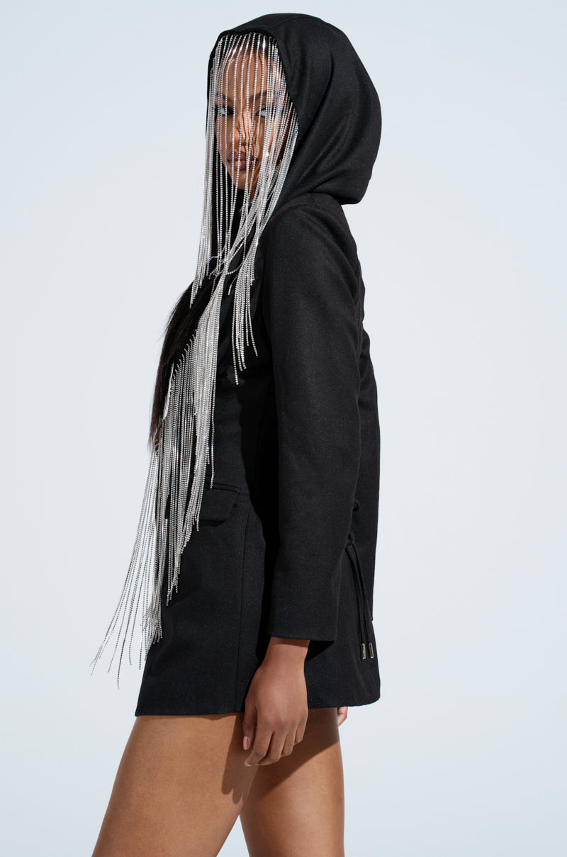 A person models the DRIPPY RHINESTONE FRINGE BLAZER, featuring a black hood with long, silver fringe. The edgy style is accentuated by lace-up corset details on the sleeves against a light background.