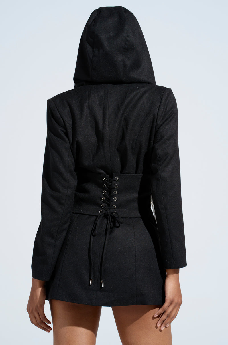 A person is shown from the back wearing a DRIPPY RHINESTONE FRINGE BLAZER, with hood up and arms relaxed by their sides. The neutral light blue background accentuates the edgy elegance of the blazer's lace-up corset detailing, cinching the waist.