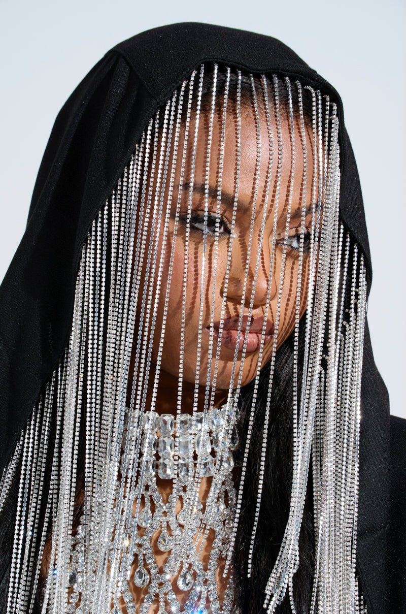 The individual is draped in a DRIPPY RHINESTONE FRINGE BLAZER, featuring a black hood and plunging neckline adorned with cascading silver chains that partially cover their face. A silver, intricately designed choker completes the look against a soft gradient of light blue and white.