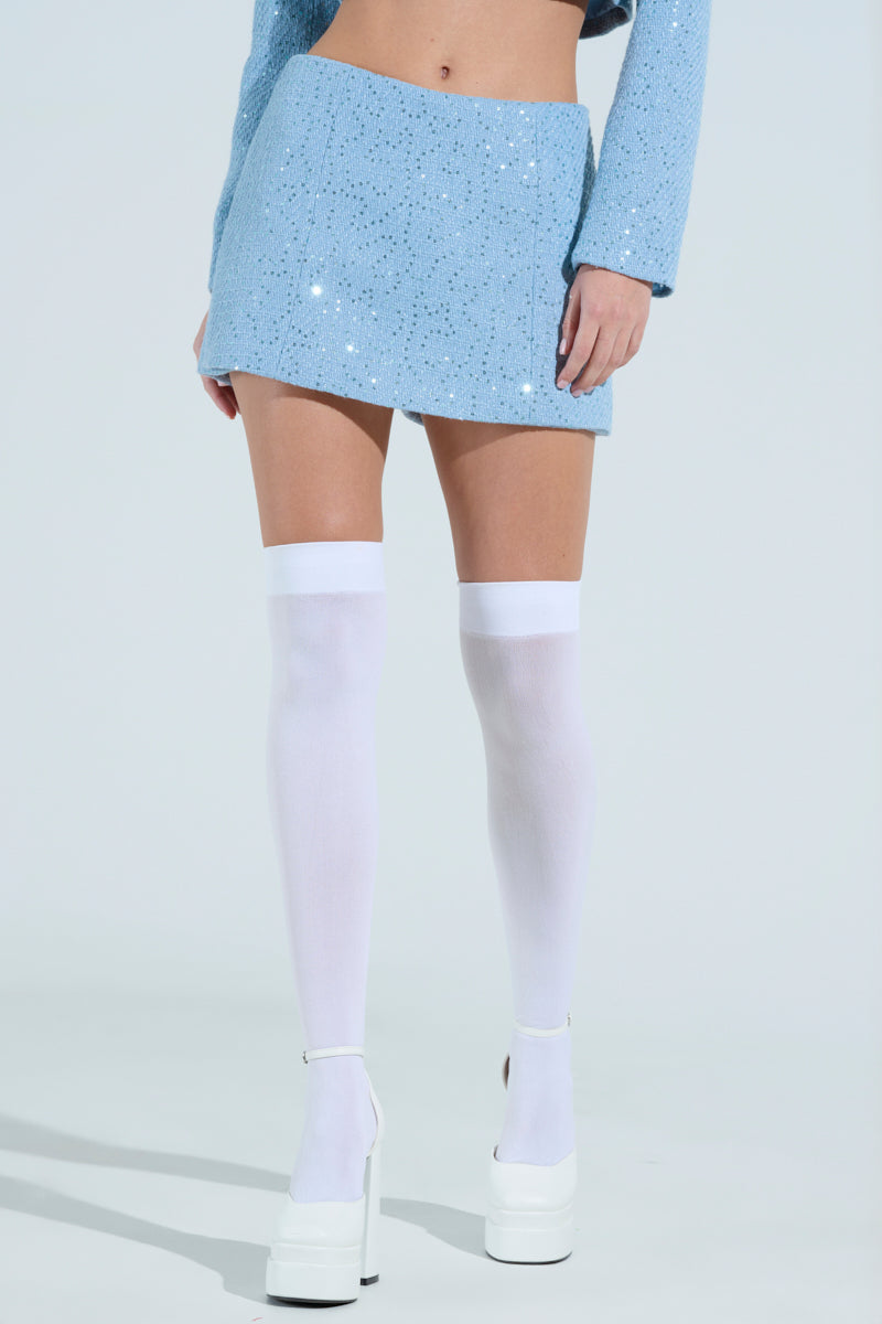 Against a pale backdrop, only the lower half of a person is visible, showcasing them in a light blue sparkly TEA FOR TWO TWEED SKORT with a high-rise silhouette and matching top, highlighted by white thigh-high stockings and chunky white platform heels.