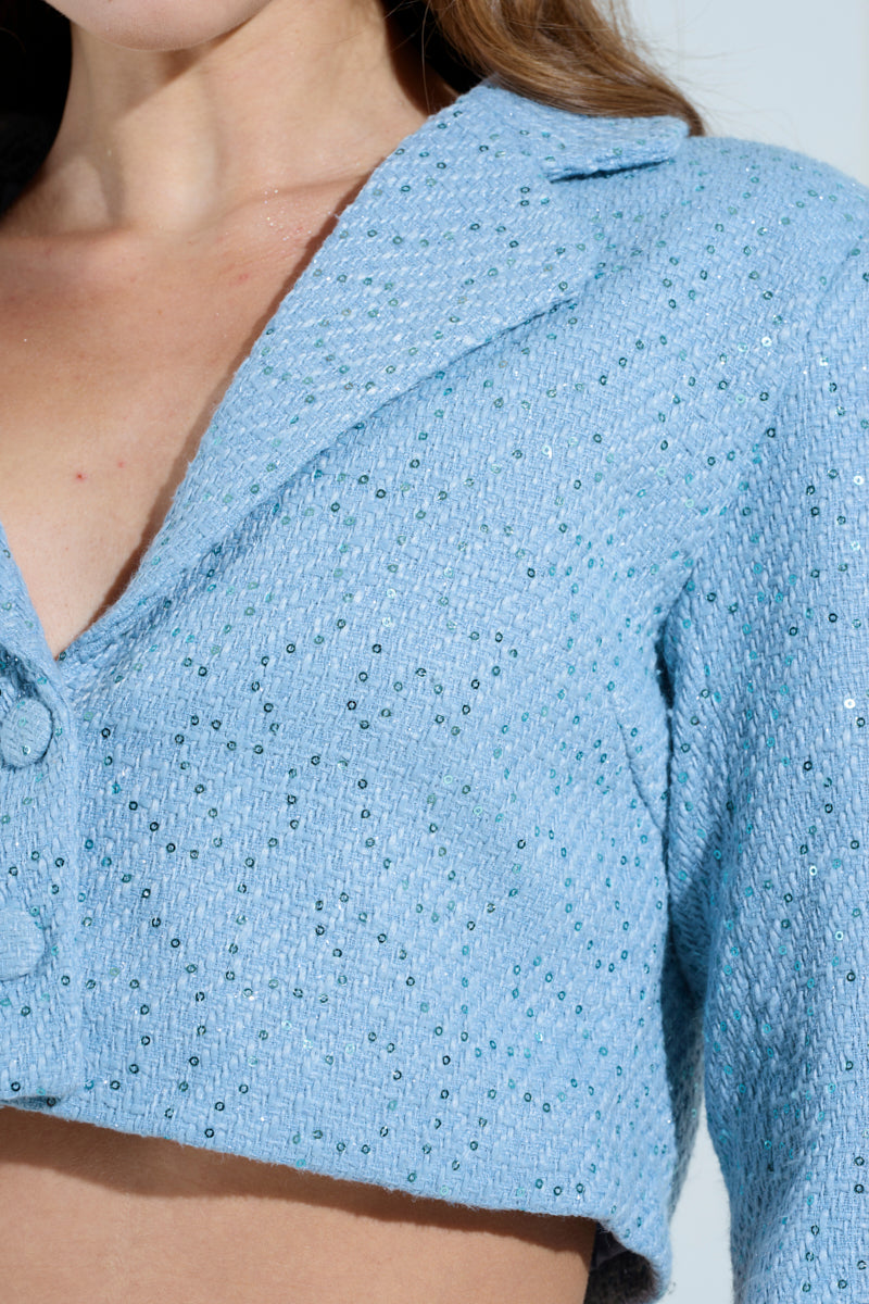 A close-up showcases a person wearing the TEA FOR TWO CROPPED TWEED BLAZER in powder blue, adorned with tiny sequins. The blazer's subtle texture and buttoned front shimmer under the light, focusing on the upper torso to highlight its exquisite details.