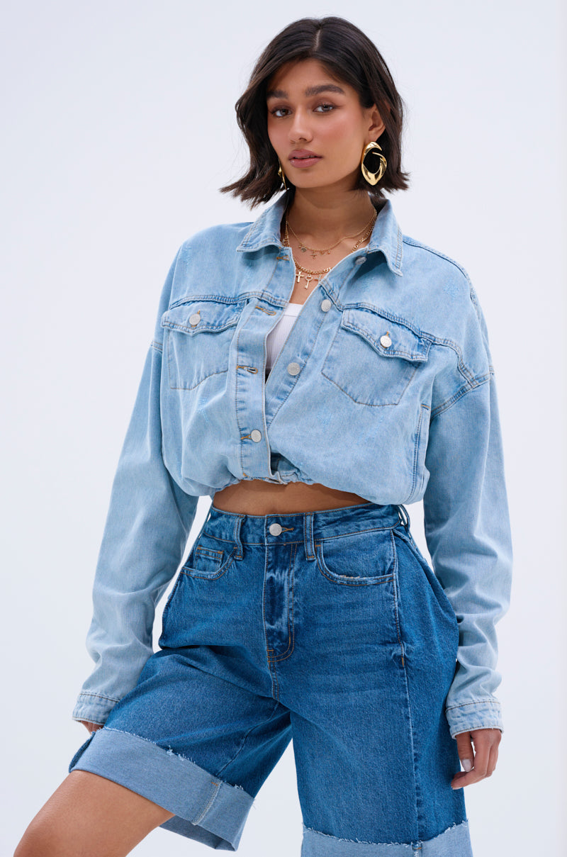 A woman with shoulder-length dark hair styles the IGGY ADJUSTABLE DENIM JACKET in light blue over a white crop top, paired with gold hoop earrings, layered necklaces, and high-waisted rolled-hem shorts for a chic streetwear look against a plain backdrop.