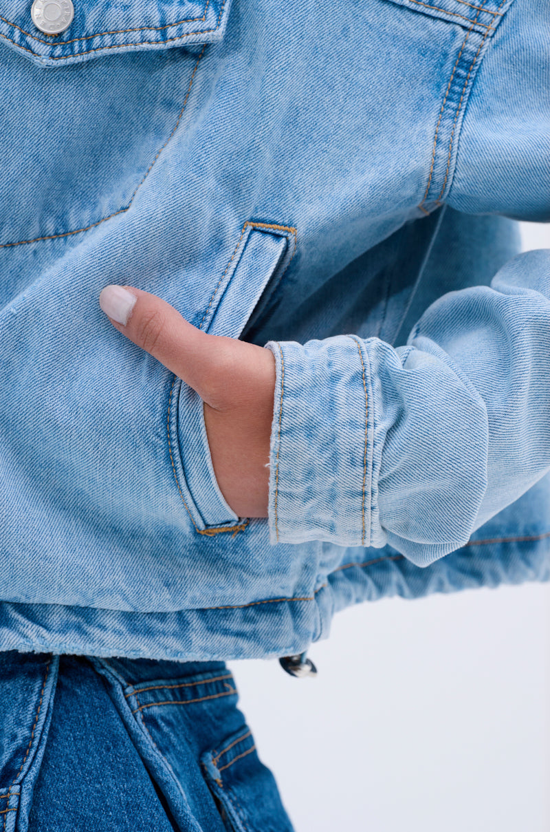 Someone wears the IGGY ADJUSTABLE DENIM JACKET in light blue vintage denim, standing with their left hand casually tucked into the pocket and the sleeve slightly bunched at the wrist, against a plain, light-colored background.