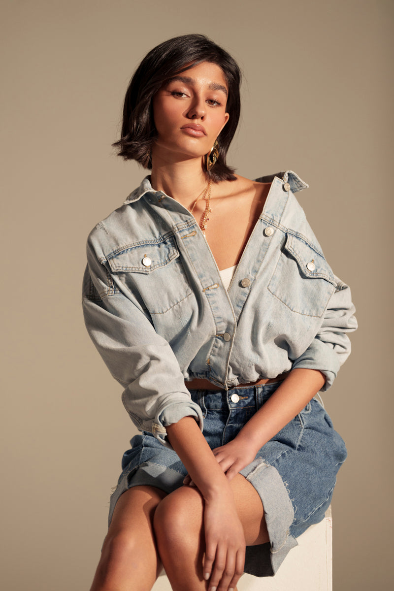 A woman with short dark hair sits on a stool, wearing the IGGY ADJUSTABLE DENIM JACKET over a white top and denim skirt. She gazes confidently at the camera, exuding vintage style against a plain neutral background.