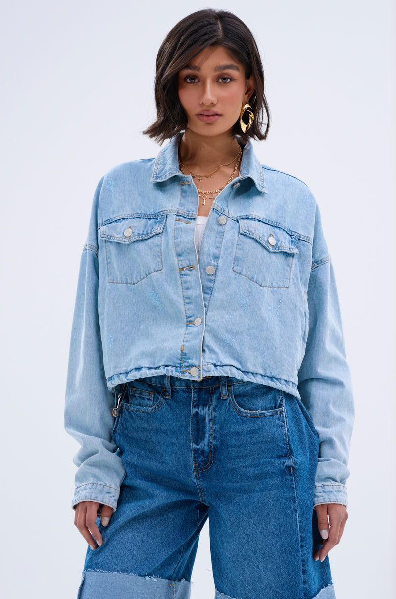 A woman with short dark hair models a vintage streetwear look in the IGGY ADJUSTABLE DENIM JACKET, cropped light blue over a white top and high-waisted patchwork jeans, accessorized with layered necklaces and large hoop earrings against a plain background.