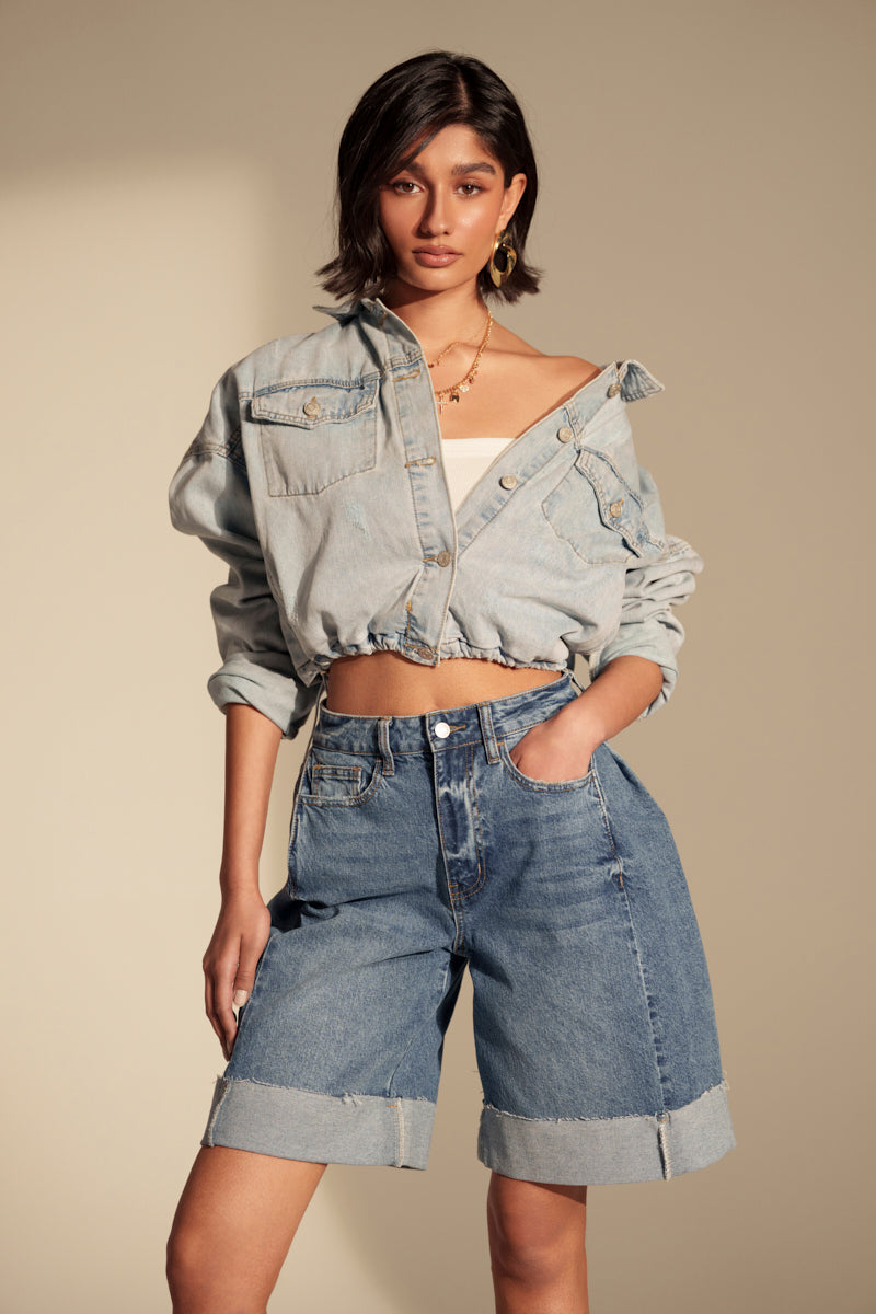 A woman exudes vintage style, wearing the IGGY ADJUSTABLE DENIM JACKET over a white top and knee-length high-waisted shorts. Gold earrings and loose waves complete her chic streetwear look as she poses confidently with one hand in her pocket.