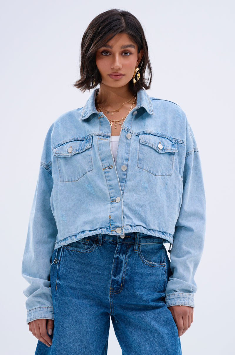 A woman with chin-length dark hair wears the IGGY ADJUSTABLE DENIM JACKET in light blue over a white top and high-waisted blue jeans, creating a subtle vintage look. She stands facing forward, expression neutral, against a plain background.