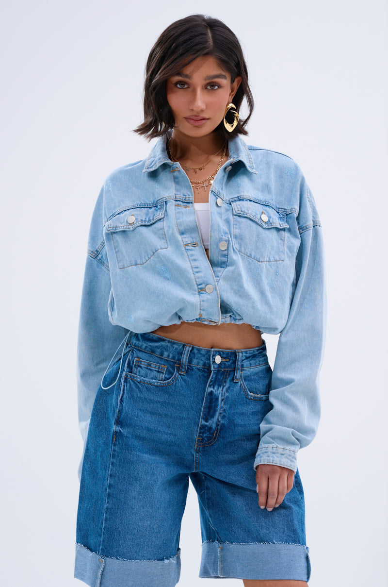 A woman with short brown hair wears the IGGY ADJUSTABLE DENIM JACKET over a white top and high-waisted cuffed shorts, finished with gold hoop earrings and layered necklaces for a vintage look against a simple light background.