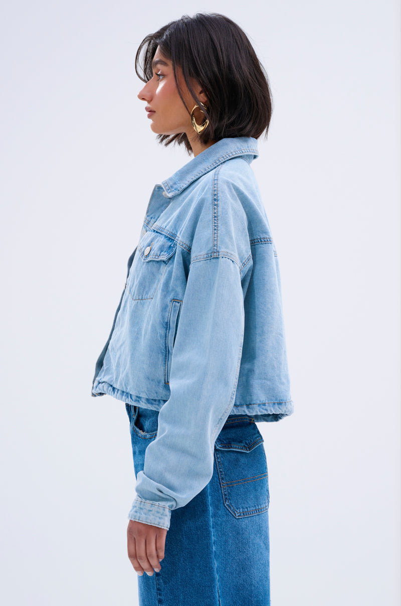 A woman with short dark hair and large hoop earrings stands in profile against a plain light background, wearing the IGGY ADJUSTABLE DENIM JACKET and blue jeans for a vintage-inspired denim-on-denim streetwear look.
