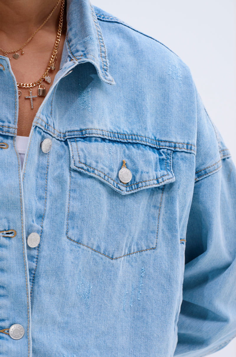 Close-up of a person wearing the IGGY ADJUSTABLE DENIM JACKET in vintage light blue, featuring metal buttons and a chest pocket, layered over a white top and accessorized with gold cross pendant necklaces.