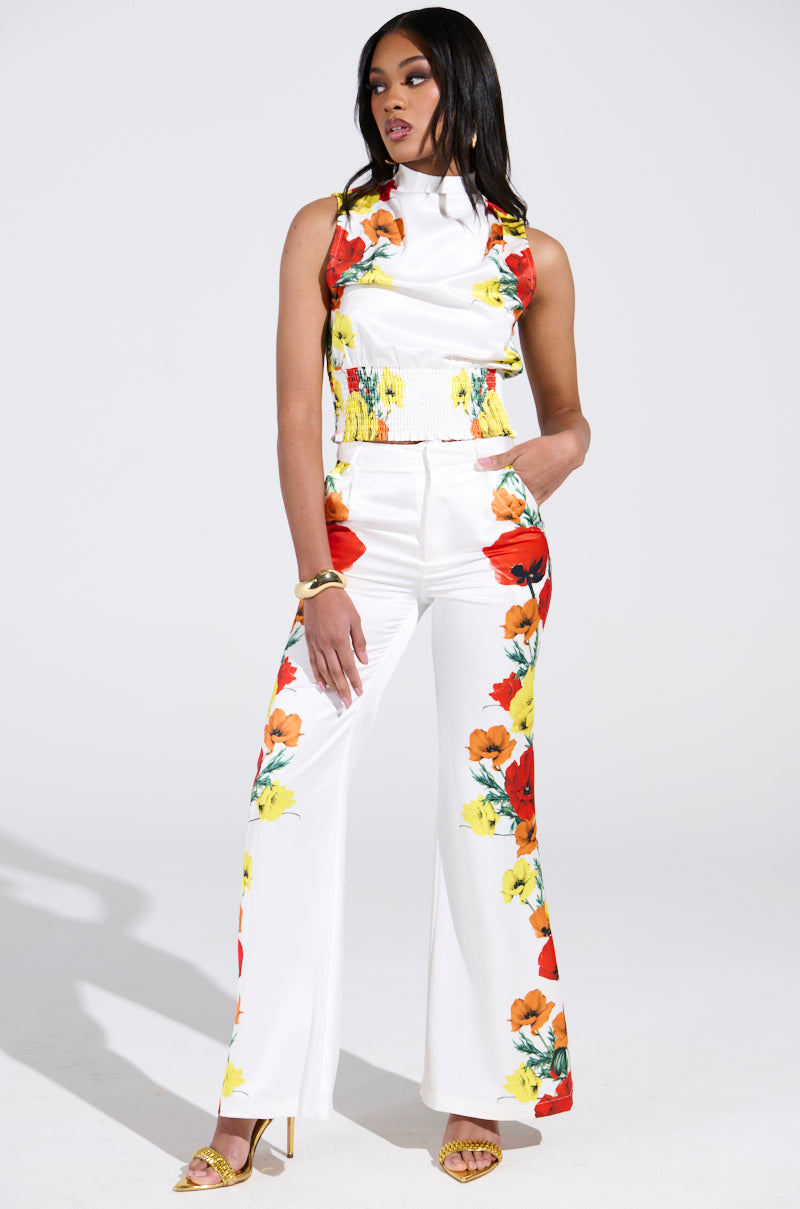 A woman confidently models white floral satin trousers, vibrant with red, yellow, and orange flowers. She pairs them with a matching sleeveless top and gold open-toe heels, one hand on her hip against a plain backdrop.