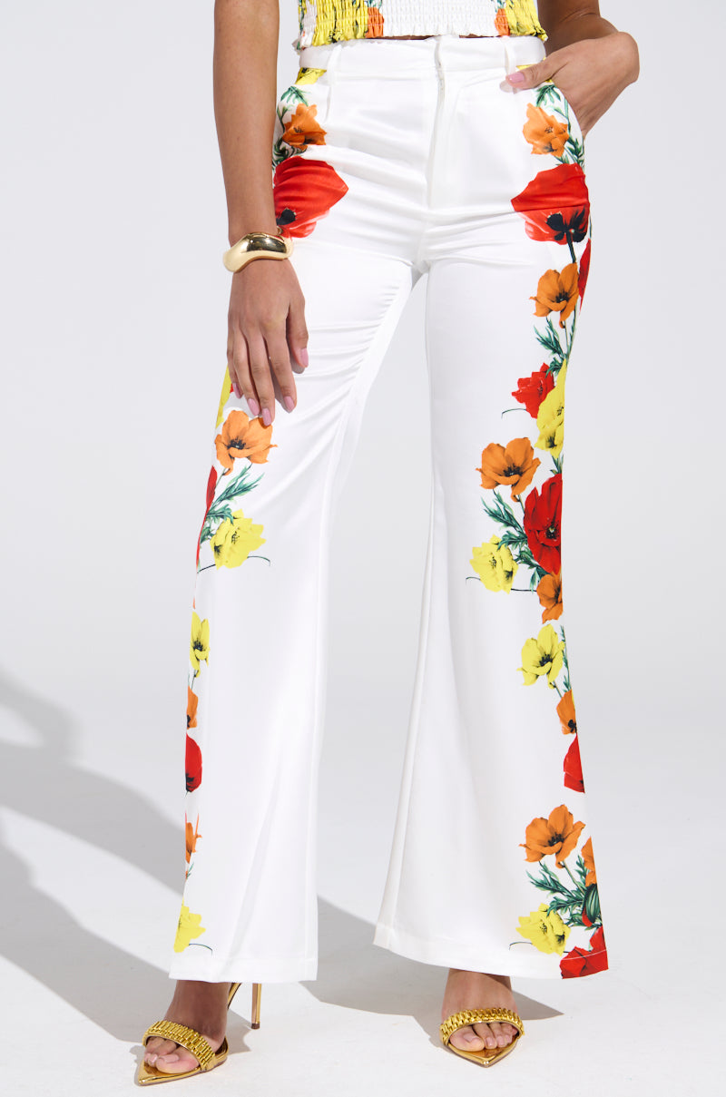 Someone poses confidently in MIND MY OWN FLORAL SATIN TROUSERS, featuring a photorealistic floral print with vibrant red, orange, and yellow blossoms. They complement the look with yellow high-heeled shoes and a gold bracelet.