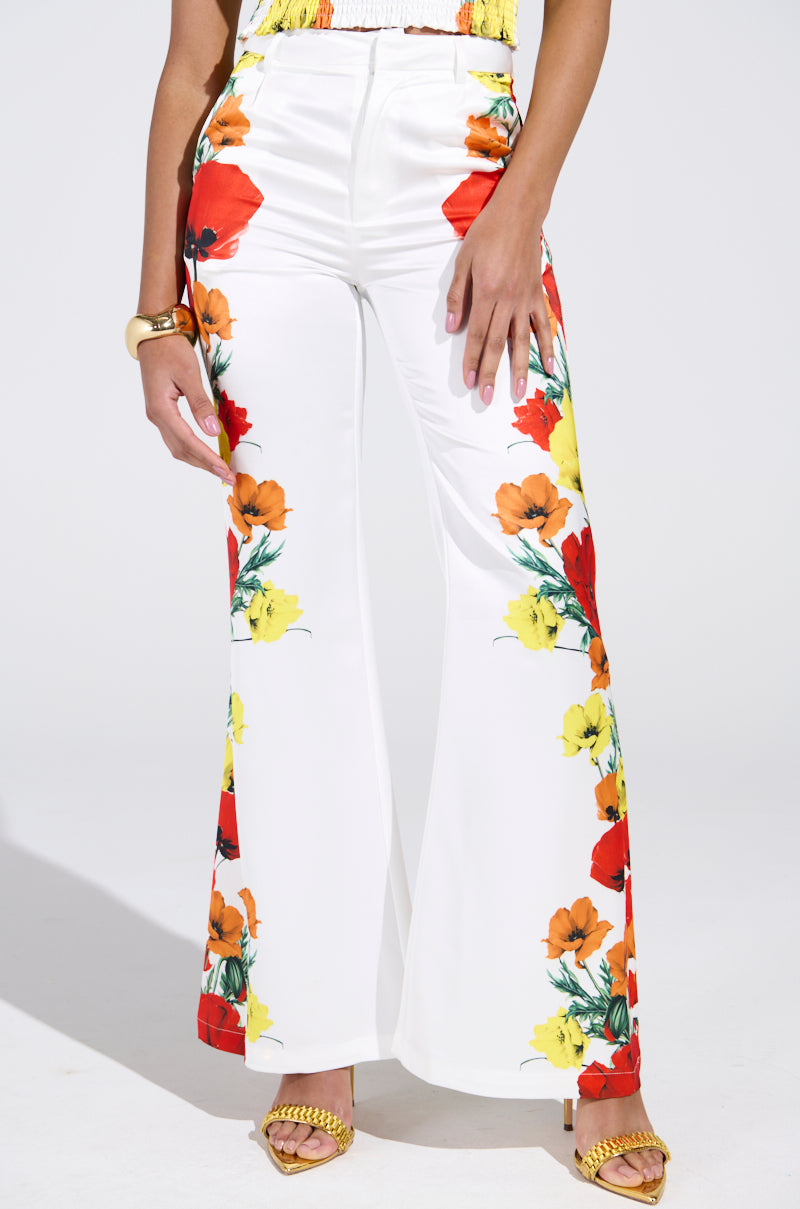 Wearing MIND MY OWN FLORAL SATIN TROUSERS with vibrant red, yellow, and orange side patterns, the person has a hand on their hip, paired with mustard-yellow high-heeled sandals and a gold bracelet.