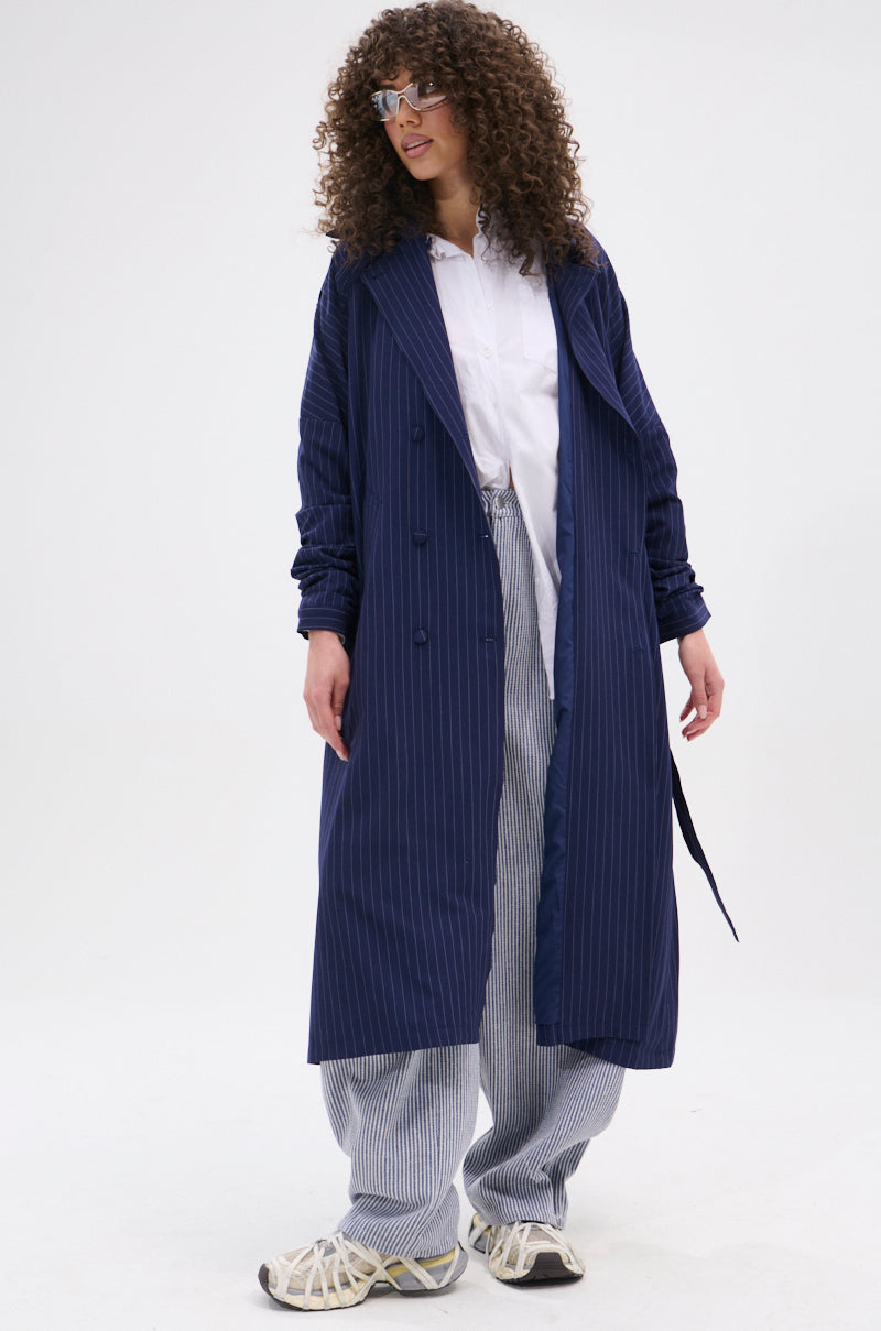 A person with curly hair and glasses wears the HARLEM PINSTRIPE TRENCH, a white shirt, loose striped pants, and white sneakers while standing against a plain white background.