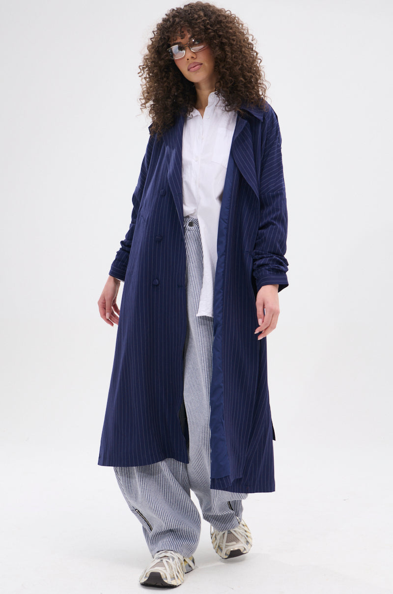 A woman with curly hair and sunglasses models the HARLEM PINSTRIPE TRENCH in a longline silhouette, styled with a white button-up shirt, loose striped pants, and white sneakers against a plain white background.