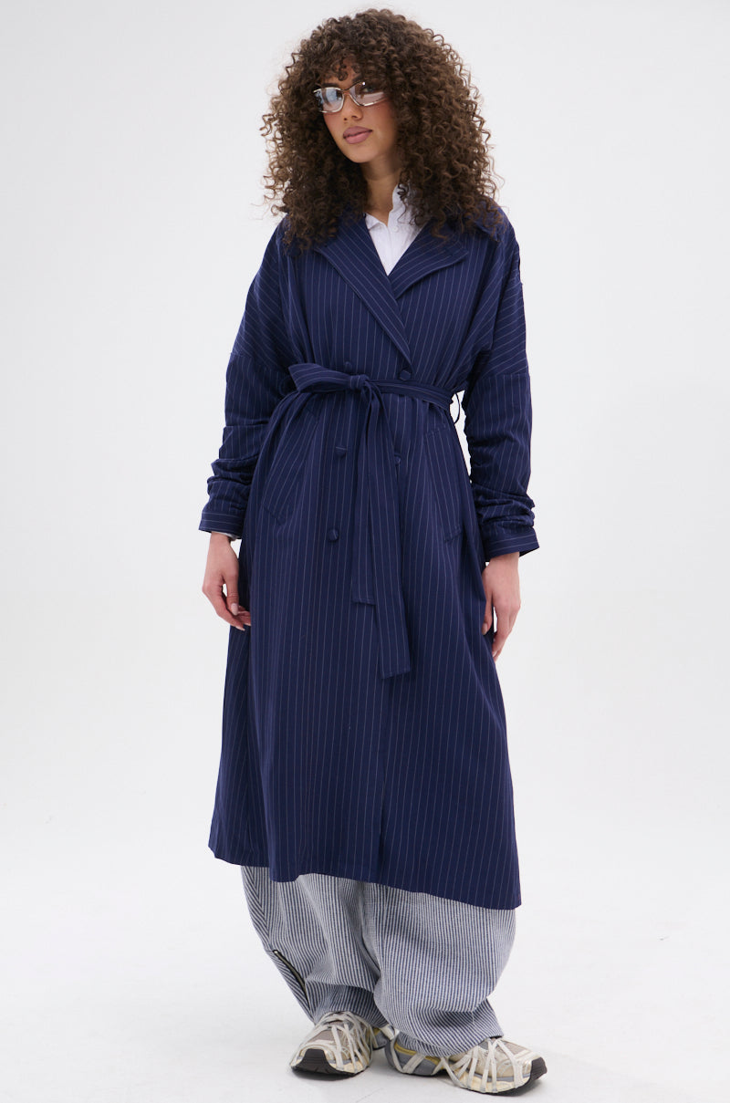 A woman with curly hair and glasses wears the HARLEM PINSTRIPE TRENCH, styled with white and blue wide-leg pants and light sneakers, standing against a plain white background.