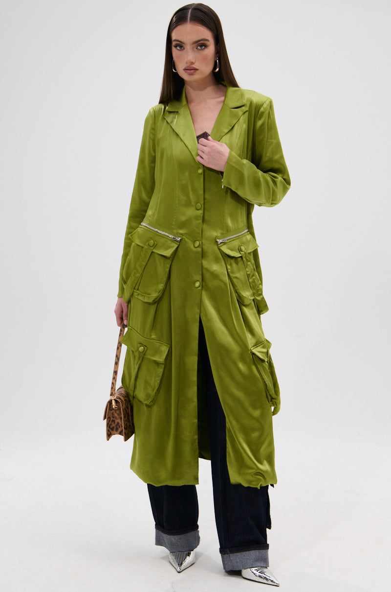 A woman stands against a white background wearing the OUTTA TIME SATIN CARGO TRENCH in lime green over dark pants with silver pointed shoes and a small leopard print bag, creating an elegant look.