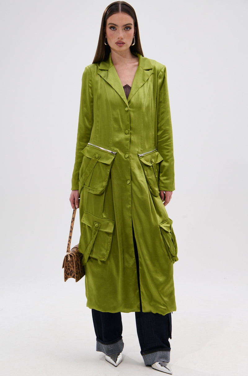 A woman in an elegant ensemble wears the OUTTA TIME SATIN CARGO TRENCH with large pockets, dark pants, and silver pointed shoes, holding a small animal-print handbag against a plain white background.
