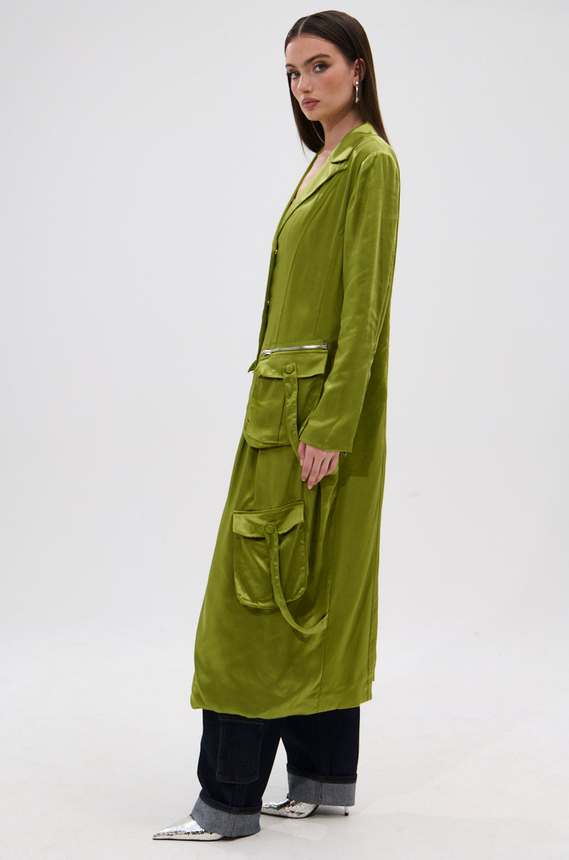 A woman stands sideways in the OUTTA TIME SATIN CARGO TRENCH from AKIRA Label, styled with dark wide-leg jeans and pointed silver shoes, set against a plain light background.
