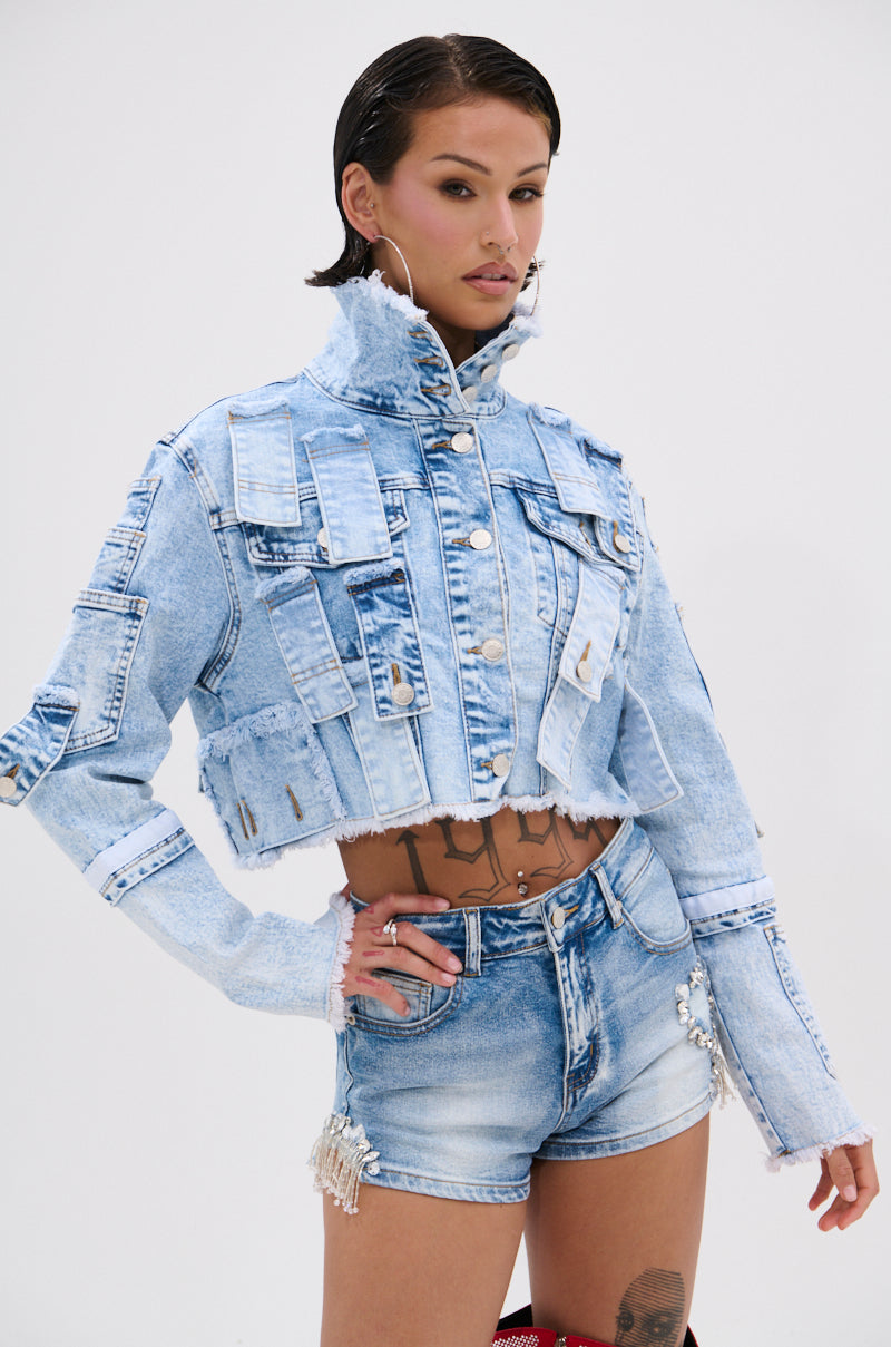 A model poses confidently in the ARIANNE PATCHWORK DENIM JACKET, styled with matching high-waisted shorts. The look features distressed trim, frayed edges, silver embellishments, slicked-back hair, and large hoop earrings.