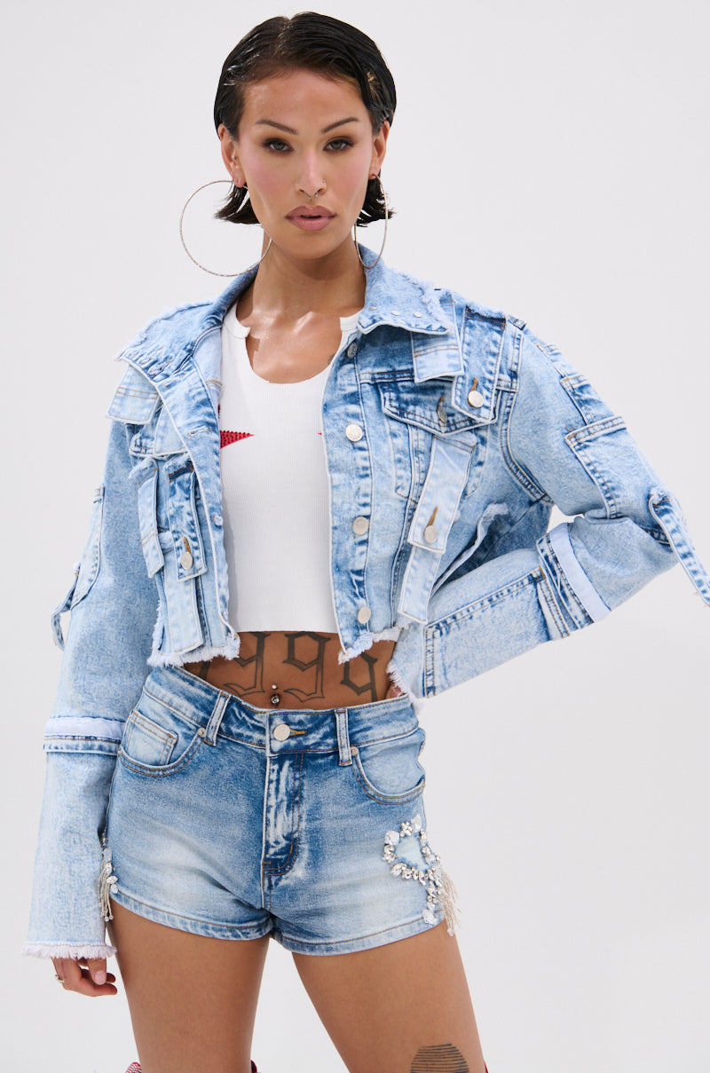 A woman poses in the ARIANNE PATCHWORK DENIM JACKET, a light blue vintage-washed style with distressed trim, paired with matching pearl-accented high-waisted shorts and a white crop top. Hoop earrings, slicked-back hair, and waist tattoos complete her look.
