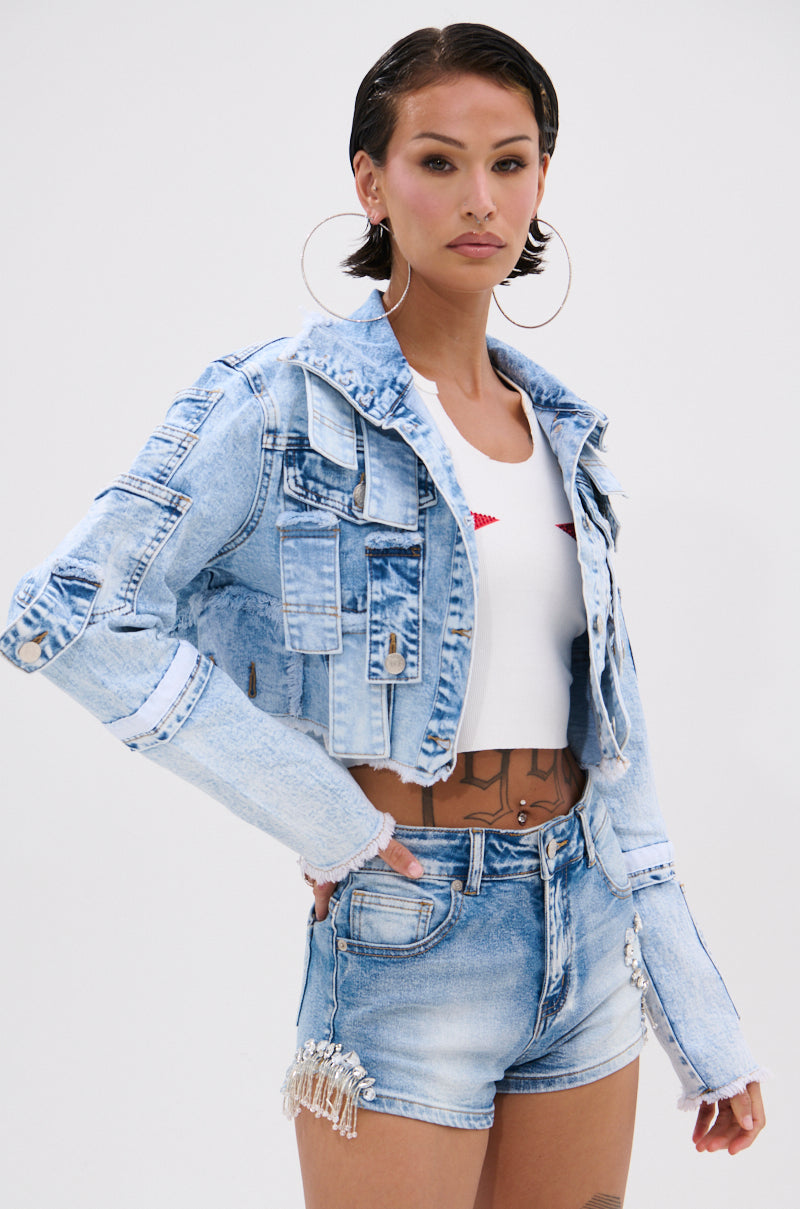 A woman with short dark hair wears the ARIANNE PATCHWORK DENIM JACKET, a white crop top, and high-waisted fringe denim shorts. She accessorizes with large hoop earrings and has visible tattoos on her torso.