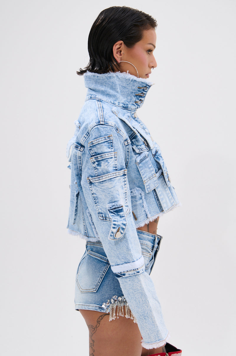 A person with short dark hair models the ARIANNE PATCHWORK DENIM JACKET, a vintage-washed cropped style with distressed trim, paired with matching high-waisted shorts. They are shown in profile against a plain white background.