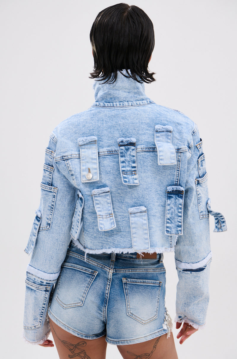 Person with short dark hair is shown from the back, wearing the ARIANNE PATCHWORK DENIM JACKET with rectangular flap details and distressed trim, paired with matching high-waisted denim shorts. Tattoos are visible on their thighs.