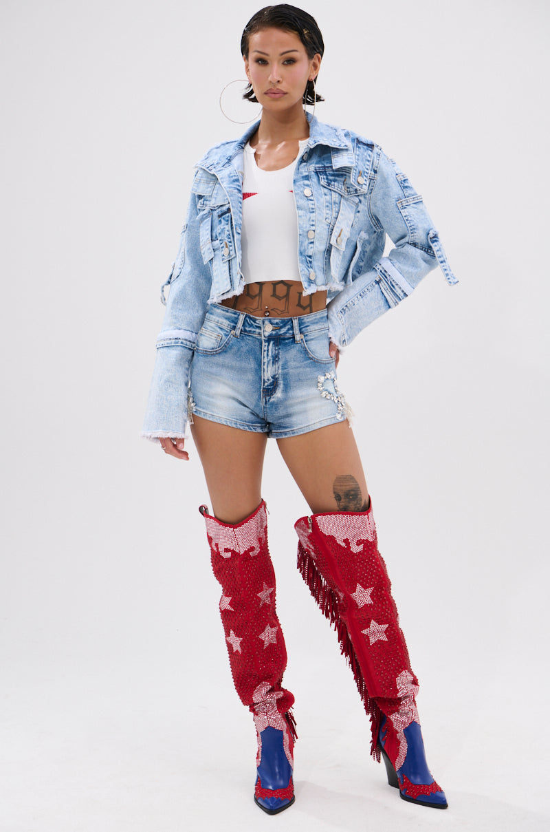 A woman poses against a white background wearing the ARIANNE PATCHWORK DENIM JACKET with distressed trim, a white crop top, denim shorts, and tall red boots with white stars and fringe. She has tattoos, hoop earrings, and a sleek hairstyle.