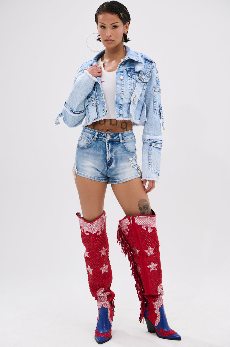 A woman exudes confidence in the ARIANNE PATCHWORK DENIM JACKET over a white top, paired with chain-detailed denim shorts and red star boots. She completes her look with tattoos, hoop earrings, and slicked-back hair.