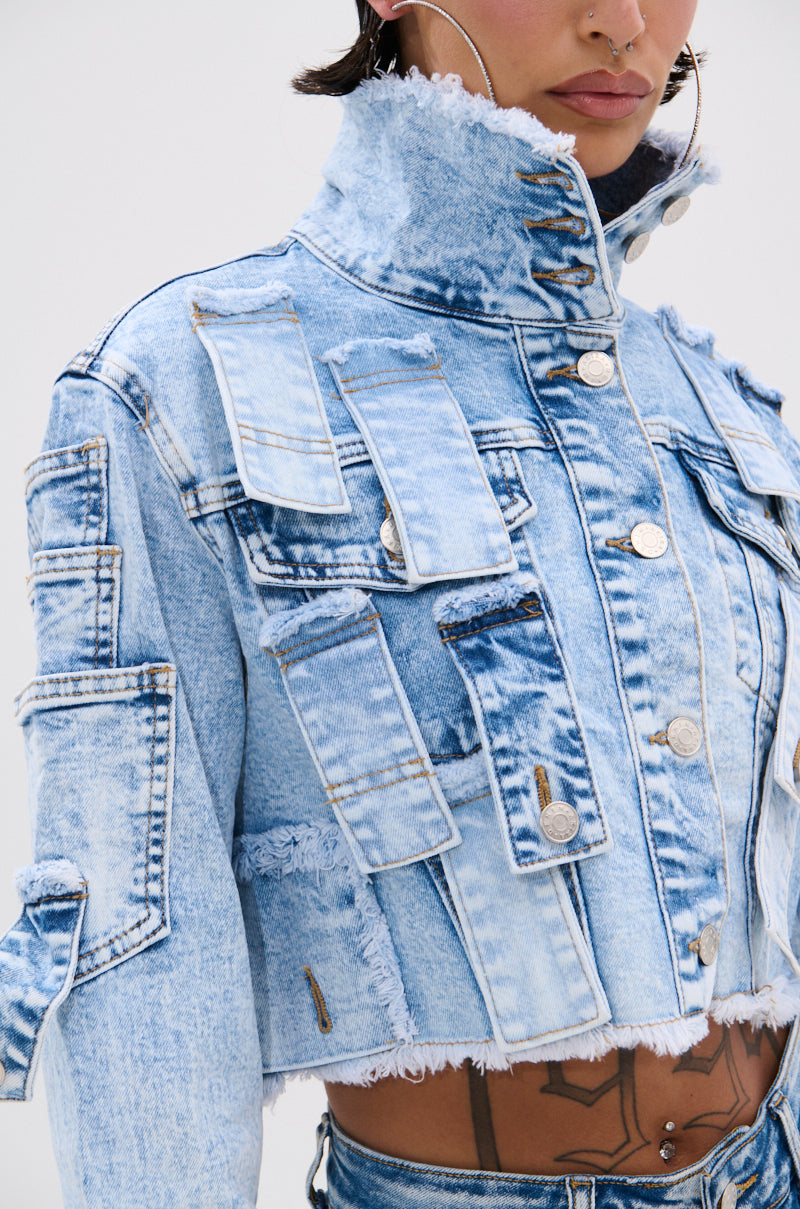 Close-up of a person wearing the ARIANNE PATCHWORK DENIM JACKET, a light blue, vintage-washed piece with multiple flap pockets and distressed trim. Tattoos are visible on their torso, and they’re accessorized with hoop nose jewelry.