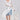 A woman stands against a light gray background wearing the BORA BORA LACE DUSTER IN WHITE over a white bustier and denim shorts, accessorized with a large woven bag and white heeled sandals.