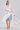 A woman stands against a light gray background wearing the BORA BORA LACE DUSTER IN WHITE over a white bustier and denim shorts, accessorized with a large woven bag and white heeled sandals.