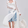 A woman stands against a light gray background wearing the BORA BORA LACE DUSTER IN WHITE over a white bustier and denim shorts, accessorized with a large woven bag and white heeled sandals.