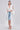 A woman wears the BORA BORA LACE DUSTER IN WHITE over a white bustier top and light blue ripped denim Bermuda shorts, styled with white heeled sandals and a textured white shoulder bag.