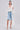A woman poses against a plain background wearing the BORA BORA LACE DUSTER IN WHITE over a white corset top and ripped denim Bermuda shorts, styled with white sandals and a textured white handbag.