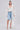 A woman wears the BORA BORA LACE DUSTER IN WHITE over a white bustier and light blue knee-length denim shorts, styled with white heeled sandals and a small white purse. Her hair is tied back as she poses against a plain background.