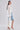 A woman stands against a plain light background wearing the BORA BORA LACE DUSTER IN WHITE over a white corset top and knee-length denim shorts, paired with white heeled sandals.