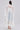 A woman stands with her back turned, wearing the BORA BORA LACE DUSTER IN WHITE over blue jeans. She carries a textured white handbag, wears white heeled sandals, and her sleek hair is tied back.