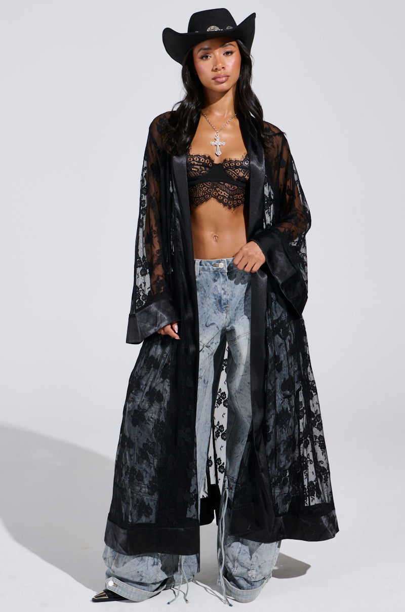 Wearing a black cowboy hat and the BORA BORA LACE DUSTER IN BLACK over a lace bralette, they exude confidence. Their festival-ready ensemble includes stonewashed jeans with white laces at the hem against a plain, light-colored backdrop that enhances their bold appearance.