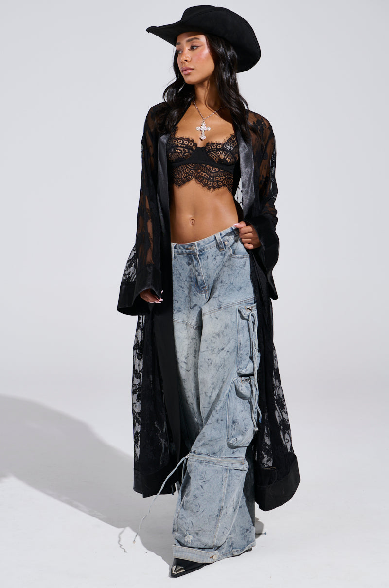 Dressed in a festival-ready outfit, the person wears a black lace bralette, the Bora Bora Lace Duster in Black, wide-leg faded jeans, black pointed boots, a silver cross necklace, and a black cowboy hat against a plain background.