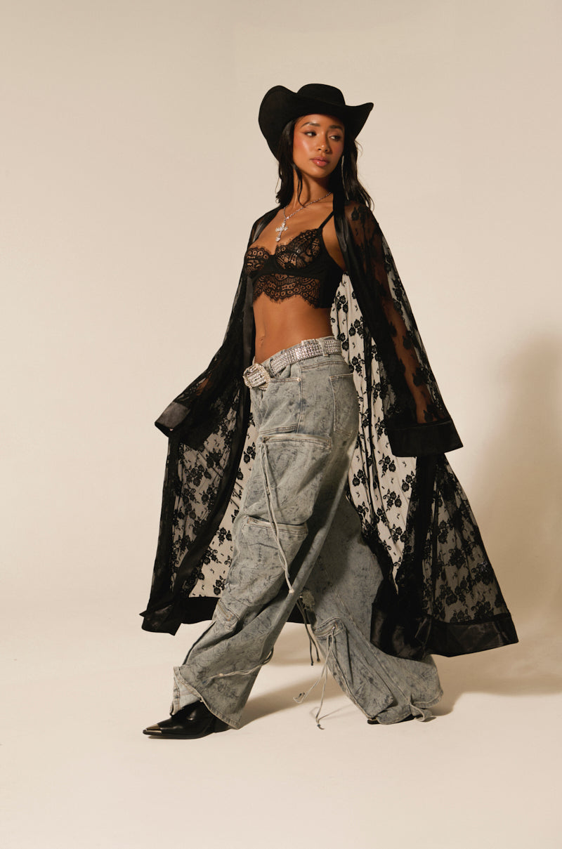 A person in a black cowboy hat, black lace top, and the Bora Bora Lace Duster strides confidently in wide-leg jeans and black cowboy boots. Their silver belt adds a striking touch, completing this bold festival outfit against a plain neutral background.