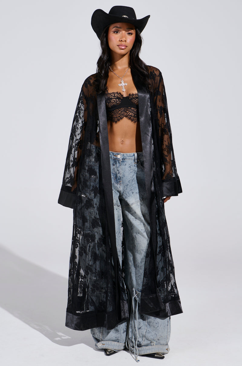 Wearing a black Bora Bora Lace Duster with a matching lace bralette and gray wide-legged pants, they embody a festival vibe. Their long dark hair flows under a black cowboy hat, while layered necklaces complete the look against a plain background.