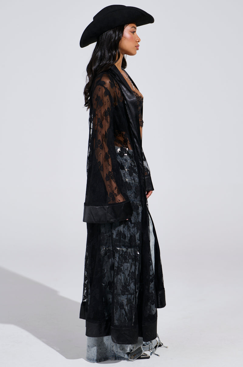 A person in a black wide-brimmed hat wears the BORA BORA LACE DUSTER IN BLACK with floral patterns over a black outfit. They stand in profile against a plain background, long dark hair cascading—a festival-ready look.